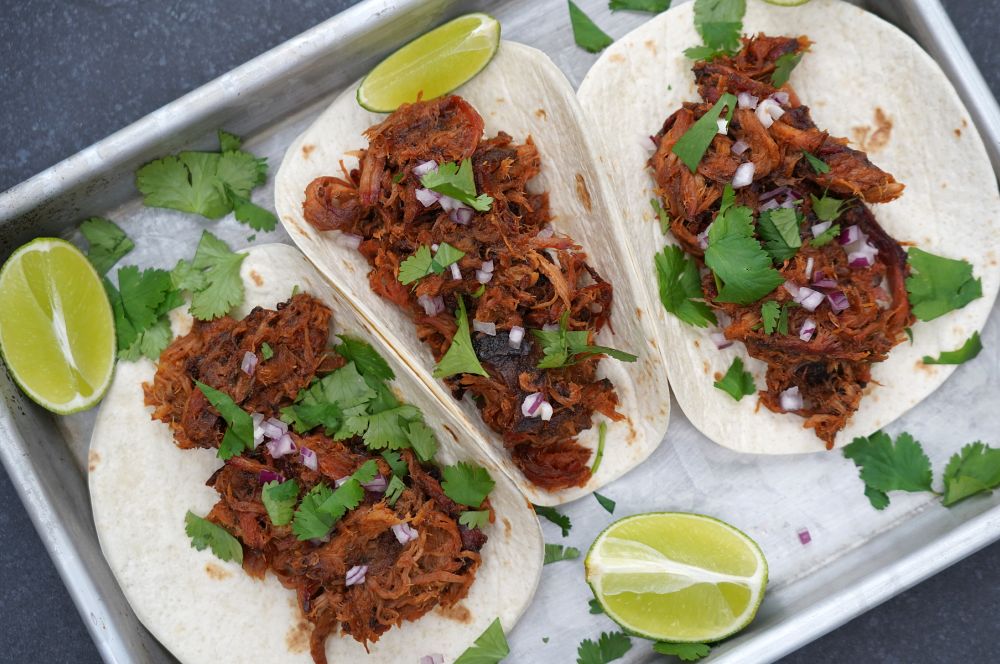 Drei Pulled-Pork-Tacos auf Metallblech mit Koriander, roten Zwiebelringen und Limettenspalten