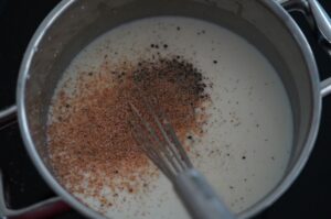 Béchamelsauce in Topf mit Gewürzen: Muskatnuss und schwarzer Pfeffer werden mit Whisk eingerührt