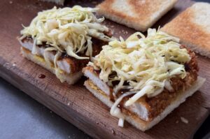 Zwei Katsu-Sando-Sandwiches auf Holzbrett mit knusprigem Schnitzel, Tonkatsu-Sauce und dünn geschnittenem Kraut auf Weizentoastbrot.