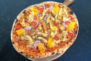 Mais-Hähnchen-Pizza mit Mozzarella, Maiskölbchen, gebratenen Hähnchenstücken, Tomaten und roten Zwiebeln auf Holzbrett