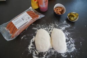 Zutaten für Pizza-Dog: zwei Pizzateigportion mit Mehl auf schwarzer Arbeitsfläche, verpackte Würstchen, Bacon, Gewürzmischung und eingelegte Gurken in Schüsseln, Getränke im Hintergrund