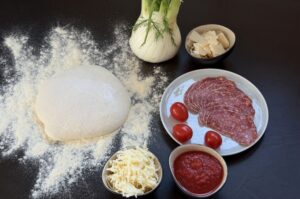 Pizzateig auf Mehl, Fenchelknolle mit Dill, geriebener Mozzarella, Fenchelsalami, Kirschtomaten und Tomatensoße für Pizza mit Fenchel und Fenchelsalami