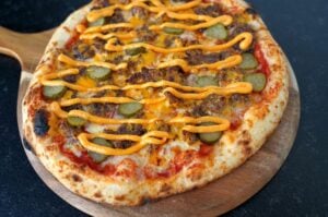 Smashburger-Pizza auf Holzbrett mit Hackfleischbelag, Cheddar, Burgersauce, Gürkchen und Zwiebelringen.