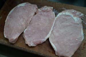 Drei rohe Schweineschnitzel mit Salz und Pfeffer gewürzt auf Holzbrett, Vorbereitung für Tonkatsu