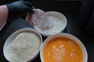 Hand mit Handschuh paniert rohes Schweineschnitzel mit Semmelbröseln, links Mehlschüssel, oben Ei, rechts fertige Panade