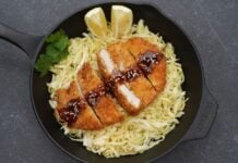 Tonkatsu in Gusseisen-Pfanne auf Gemüsenudeln angerichtet, mit Teriyaki-Glasur, Sesamkörnern und Zitronenscheiben