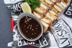 Schüssel mit dunkler Tonkatsu-Sauce mit Sesamkörnern neben gegrilltem Tonkatsu mit knuspriger Panade, Zitronenscheibe und frischer Petersilie