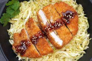 Panierte Schweineschnitzel auf Nouilles, mit glänzender Tonkatsu-Sauce und Sesam garniert, Koriander-Deko