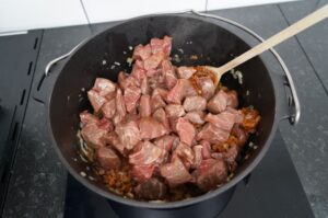 Rinderfleischwürfel werden in schwarzem Topf auf Grillfeld mit Holzlöffel gerührt, Zwiebeln angeröstet, Zubereitung Wiener Saftgulasch