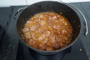 Wiener Saftgulasch in schwarzem Topf auf Grillplatte: Rinderfleischstücke in braun-roter Soße mit Zwiebeln.