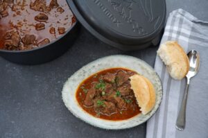 Wiener Saftgulasch in Schmorpfanne und auf Teller mit Semmel serviert, garniert mit Petersilie und Zwiebeln