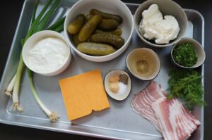 Zutaten für Gurkendip: Sauerrahm, eingelegte Gurken, Frischkäse, Bacon, Schnittlauch, Dill, Gewürze und Öl auf grauem Tablett