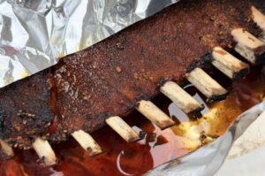 Wildschwein-Ribs mit dunkler Rindenkruste auf dem Grill, Räucherpapier dahinter, Fleisch zwischen Knochen sichtbar