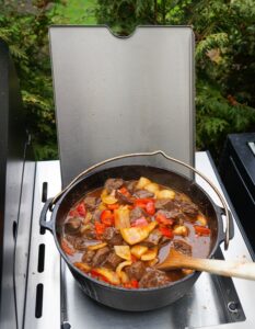 Brauhaus-Gulasch in schwarzem Dutch Oven auf Grillplatte: Rinderfleischwürfel mit Kartoffeln und roten Paprikastücken in dunkler Soße, Holzlöffel zum Umrühren