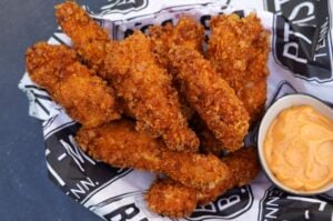 Knusprig panierte Hähnchen-Finger mit goldbrauner Kruste auf schwarzweißem Papier mit Sauce in Schälchen daneben