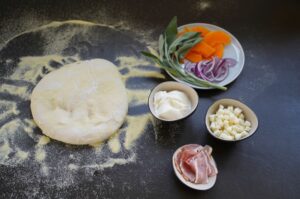 Kürbispizza-Zutaten: Pizzateig auf bemehlter Oberfläche, Kürbis und rote Zwiebeln, Salbeiblätter, Creme fraîche, Käse und Speck in Schälchen