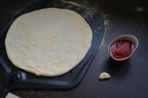 Pizzateig auf schwarzem Grillblech, daneben Schüssel mit Tomatensoße und Knoblauch