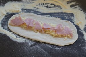 Pizzabrötchen-Teig mit Käsestreifen und Schinkenstreifen belegt, auf bemehlter schwarzer Arbeitsfläche