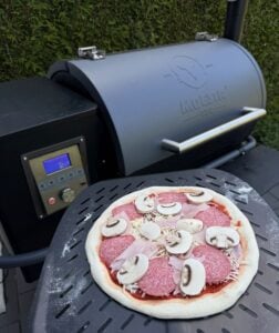 Pizza mit Salami und Pilzen auf schwarzem Grillrost eines Pelletgrills mit digitaler Temperaturanzeige im Hintergrund