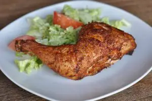 Gegrillter Hähnchenschenkel mit knuspriger, braun gebräunter Oberfläche auf weißem Teller mit grünem Salat und Tomatenscheibe