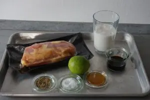 Kokos-Limetten-Hähnchenspieße 8 Zutaten für Kokos-Limetten-Hähnchenspieße auf Edelstahlplatte: Hähnchenbrust in Vakuumverpackung, grüne Limette, Kokospulver, Salz, Honig, Sojasoße und Glas mit Wasser