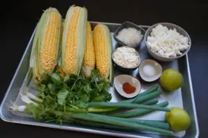 Zutaten für Esquites: vier frische Maiskolben mit Blättern, Koriander, Frühlingszwiebeln, Limonen, Maiskörner, Frischkäse, geriebener Käse, Salz und Paprikapulver auf weißem Tablett.