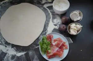 Zutaten für Feigenpizza: ausgerollter Pizzateig, Serrano-Schinkel mit Rucola, Ziegenkäse, rote Feigen und Creme fraîche auf dunklem Untergrund