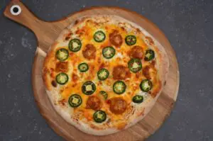 Jalapeno-Popper-Pizza auf Holzbrett: Käse, Chorizo-Stücke und grüne Jalapeño-Scheiben auf Teigfladen mit Brandflecken