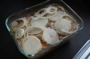 Thüringer Rostbrätel mit Zwiebeln in Glasauflaufform, mariniert mit Sauce und frischen Zwiebelringen, vor dem Grillen