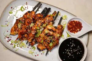 Yakitori-Spieße mit glasiertem Hähnchenfleisch, Sesam, grünem Frühlingszwiebel-Topping und Chilliflocken. Dazu Schälchen mit Sojasauce und Chili-Öl.