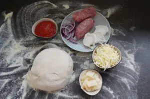 Pizzateig und Zutaten für Salsiccia-Pizza: italienische Wurstscheiben, rote Zwiebeln, Ziegenkäse, geriebener Mozzarella, Butter und Tomatensoße