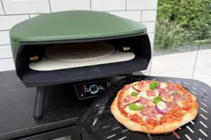 Salsiccia-Pizza mit Mozzarella, Salami und roten Zwiebeln auf Pizzaofen-Kochplatte, grüner Deckel geöffnet.