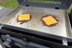 Zwei Smash-Burger mit Cheddar-Käse auf einer Grillplatte eines Urban II Pro Grills, im Hintergrund grüne Rasenfläche