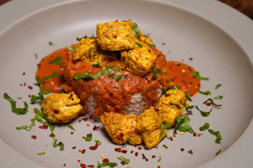 Chicken Tikka Masala mit würzigen Hühnchestücken auf Cremesauce und Reisbett, garniert mit grünen Kräutern und Chilischoten