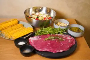 Zutaten für Flanksteak mit Pesto-Nusskruste: Rohes Flanksteak mit Rosmarin, Maiskolben, Rucola, Pinienkerne, Pestogewürz, Salz und Pfeffer, sowie Gemüsewürfel in Edelstahlschale