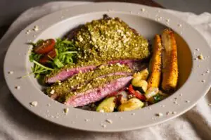 Gegrilltes Flank Steak mit grüner Pesto-Nusskruste, medium-rare geschnitten, dazu gegrillte Würstchen und Gemüse mit Kartoffeln auf grauem Teller