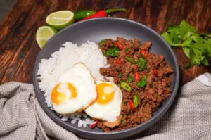 Pad Kra Pao mit zwei Spiegeleiern, Hackfleisch mit grünen und roten Chilis, dazu Jasminreis in grauer Schale auf Holzbrett