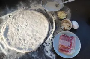 Pizzateig mit Mehl auf schwarzem Untergrund, daneben Zutaten: Schüssel mit Creme, Speck, geriebener Käse, gehackte Zwiebeln, Ei
