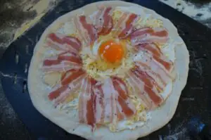 Pizza Carbonara mit Spiegelei auf Grillplatte: Teig mit Speckstreifen in Blütenform arrangiert, in der Mitte rohes Eigelb, gekochte Nudeln, schwarzer Pfeffer, auf dunkler Grillplatte.