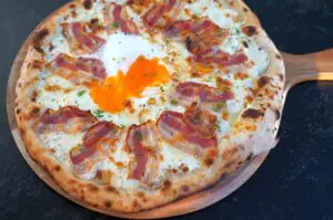 Pizza Carbonara mit Spiegelei, belegt mit Pancetta, Mozzarella und flüssigem Eigelb, auf Holzbrett