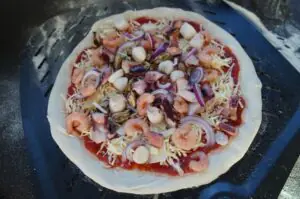 Meeresfrüchte-Pizza mit Garnelen, Muscheln, Tintenfisch, geriebenem Käse, roten Zwiebeln und Tomatensoße auf schwarzem Grillblech
