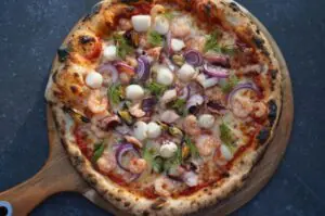 Pizza Frutti di Mare mit Garnelen, Muscheln, roten Zwiebeln, Mozzarella und Dill auf Holzbrett