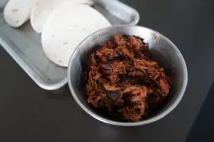 Geschreddertes Pulled Pork in einer Metallschale mit Tortillas im Hintergrund zur Zubereitung von Pulled Pork Tacos