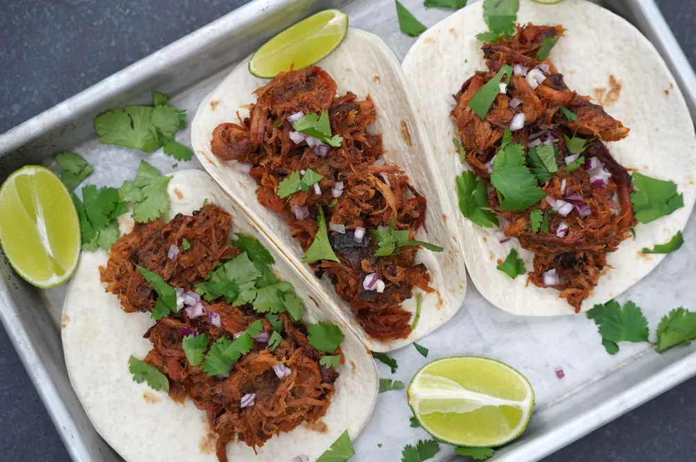 Drei Pulled-Pork-Tacos auf Metallblech mit Koriander, roten Zwiebelringen und Limettenspalten