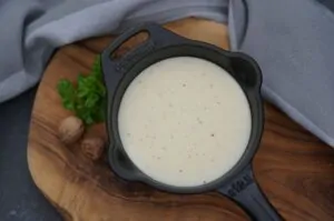 Béchamel-Sauce in schwarzem Gusseisen-Topf auf Holzbrett, garniert mit Petersilie und Muskatnuss