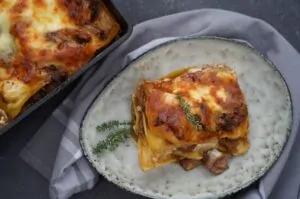 Gulasch-Lasagne - Das herzhaft überbackene Schmorgericht 16 Gulasch-Lasagne auf Teller mit Thymian garniert, daneben Auflaufform mit restlicher Lasagne, goldbraune Käsekruste