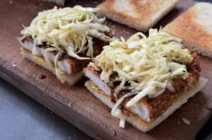 Zwei Katsu-Sando-Sandwiches auf Holzbrett mit knusprigem Schnitzel, Tonkatsu-Sauce und dünn geschnittenem Kraut auf Weizentoastbrot.