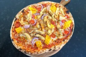 Maishähnchen-Pizza mit Pollo Fino und Mais 10 Mais-Hähnchen-Pizza mit Mozzarella, Maiskölbchen, gebratenen Hähnchenstücken, Tomaten und roten Zwiebeln auf Holzbrett