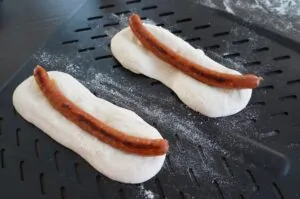 Zwei Pizza-Dogs mit gegrillter Bratwurst in Pizzateig auf schwarzem Grillrost, mit Mehls­taubung
