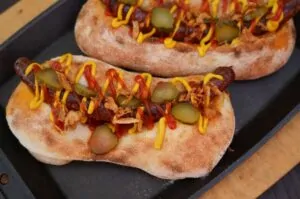Zwei Pizza-Dogs mit Wursteinlage, Senf und roten Peperonistreifen, sowie eingelegte Gurken auf schwarzem Grillblech